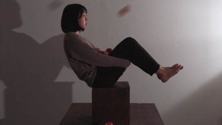 Video still showing a young adult holding their knees to create a sort of basket with their body while balancing on a box. Apples are thrown into their lap by someone off-camera.