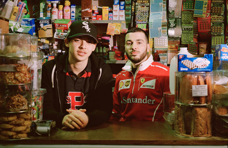 Two male adults standing behind the counter of a convenience store, looking straight ahead and surrounded by packaged food.