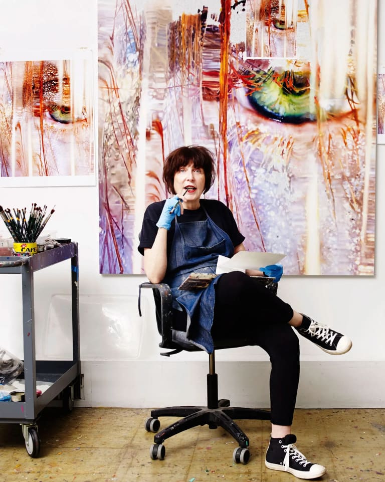 A woman sits on a rolling chair in a studio in front of a large brightly colored painting 