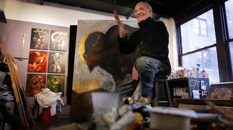 An older gentleman, sitting on a stool, gestures towards the camera as an array of paintings and art supplies can be seen in the background.