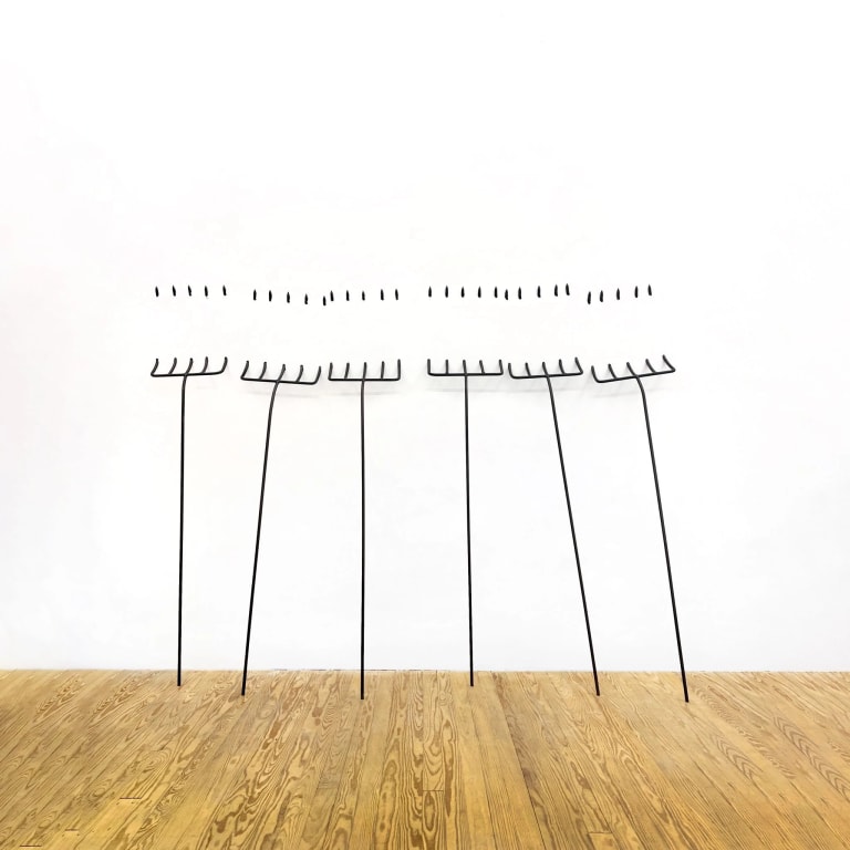 Sculptural installation in a white gallery space consisting of six thin black pitchforks leaning against a wall. The tines of the pitchforks penetrate the wall, and their points seem to emerge from the same wall about a foot above where they enter. 