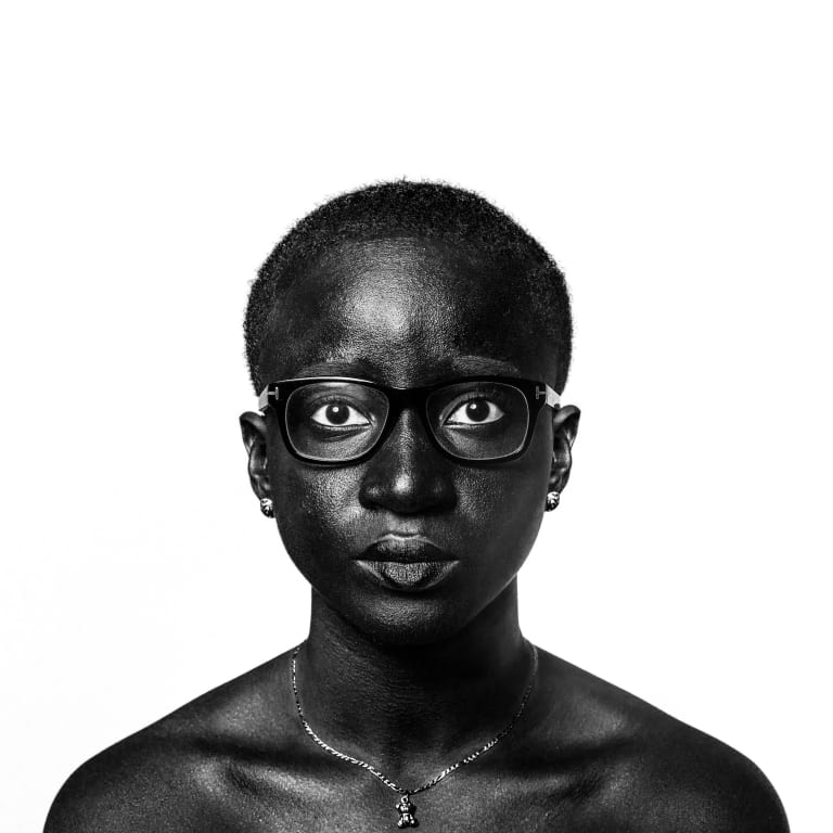 A studio portrait of a black woman from the shoulders up. She is wearing black glasses, a silver necklace, and silver stud earrings. She is staring at the camera with a neutral expression. She has very short, close-cropped hair. The background is white.