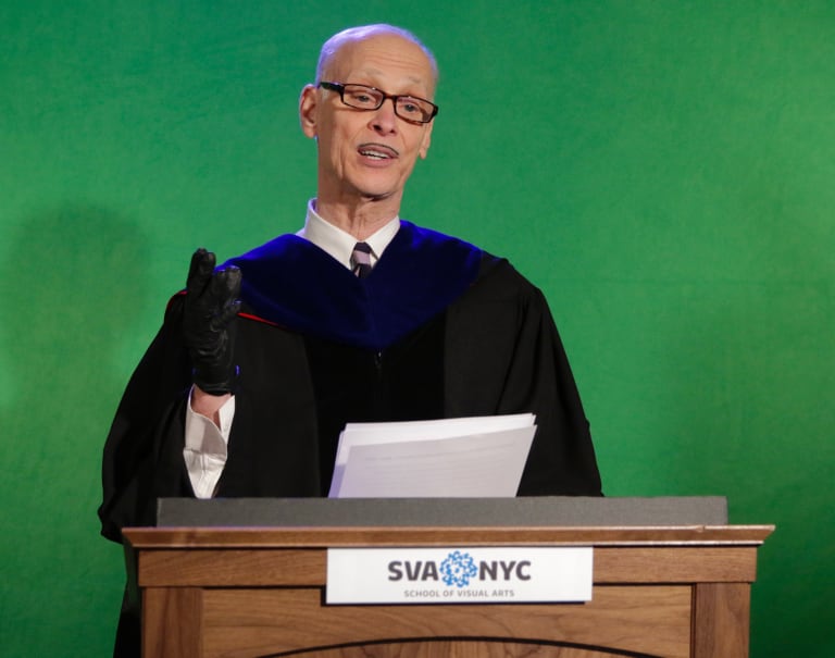 A photo of a man in a tie and graduation gown, standing at a podium set in front of a green screen.