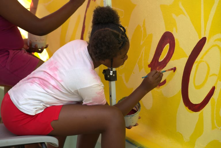 A person in a t-shirt in shorts sits on a chair, crouching to paint a yellow wall with red paint