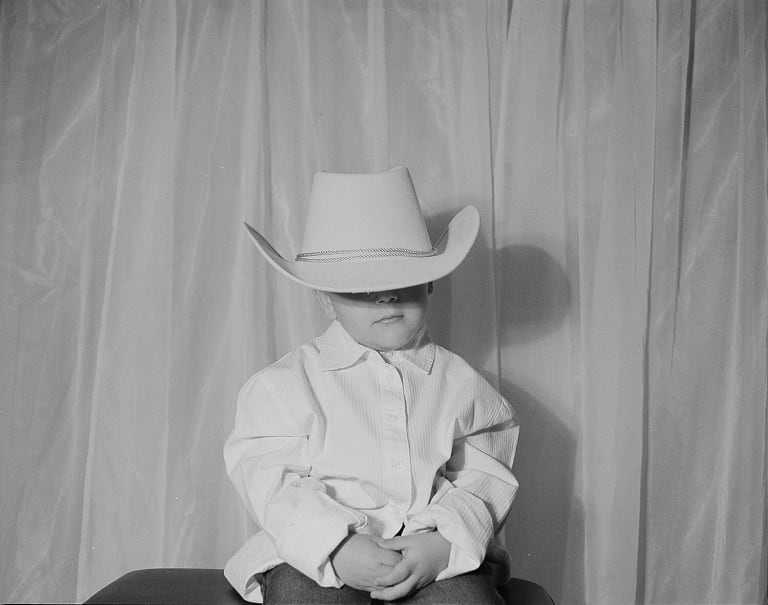 Black and white photo of a child wearing a cowboy hat that is too big for them, obscuring their face.
