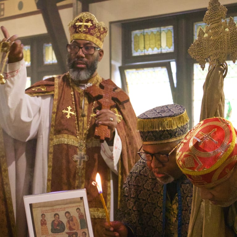 Three men are in a church wearing Ethiopian Orthodox priest garments, which are boldly colored with gold accents. One man stands and swings a gold incense burner; the other two hang their heads and bow.