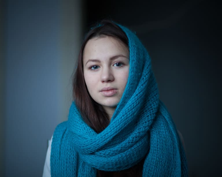 A horizontal photographic portrait of a young woman with blue eyes looking at the viewer. The image has a cool tonality and the woman’s head is loosely wrapped in a blue woolen scarf reveling some of her hair.
