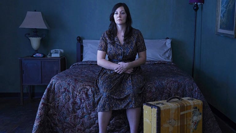 A woman sitting on the bed in a dark room with a suitcase near her feet.