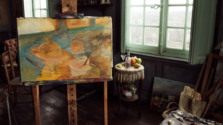 Inside a painting studio with a painting on an easel to the left, a still left in the middle back, and oil paint and canvas to the lower right.