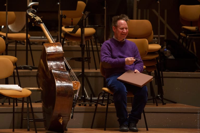 A photograph of Peter Sellars sitting in a chair in an orchestra room, smiling at something beyond the camera.