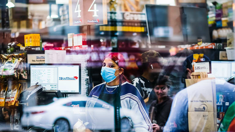 In this photo, a masked cashier looks through the window of a supermarket, while the street outside is reflected in the window surface.