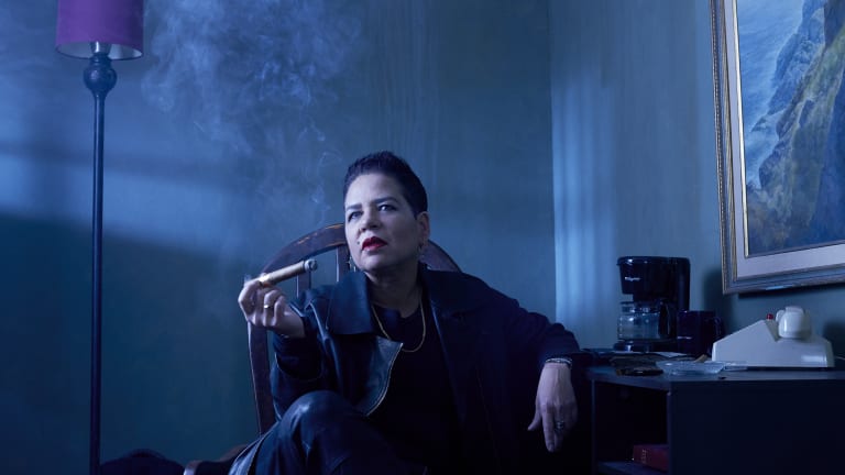 Woman sitting at a desk in a blue lit space smoking a cigar.