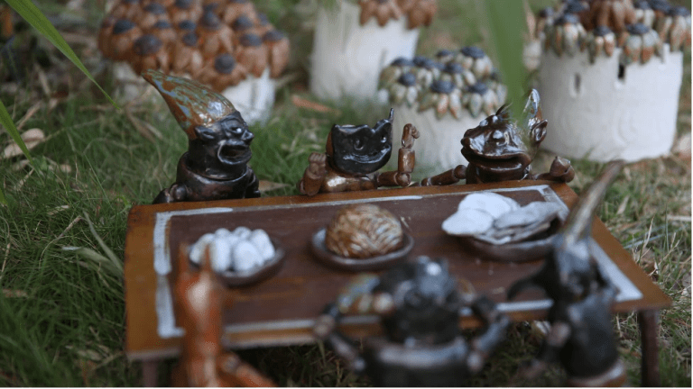 Video still showing six miniature ceramic figures sitting around a table set in a grassy area. 