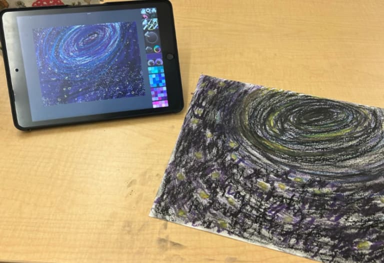 A tablet shows a digital drawing of outter space next to the paper version of the drawing.