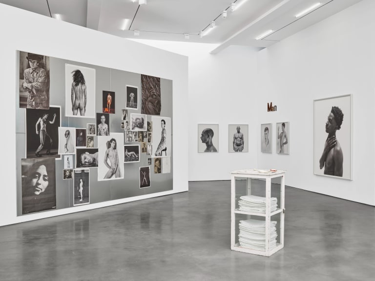 White gallery room with photographs hung in a salon style on left wall; framed portraits on right wall. Sculpture in the center of the room