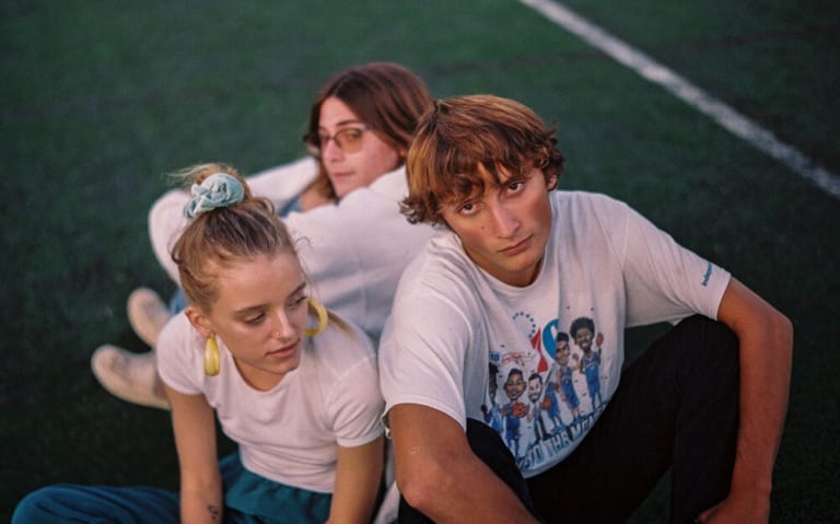 Three teens wearing white T-shirts sit on a football field in a cluster with moody looks