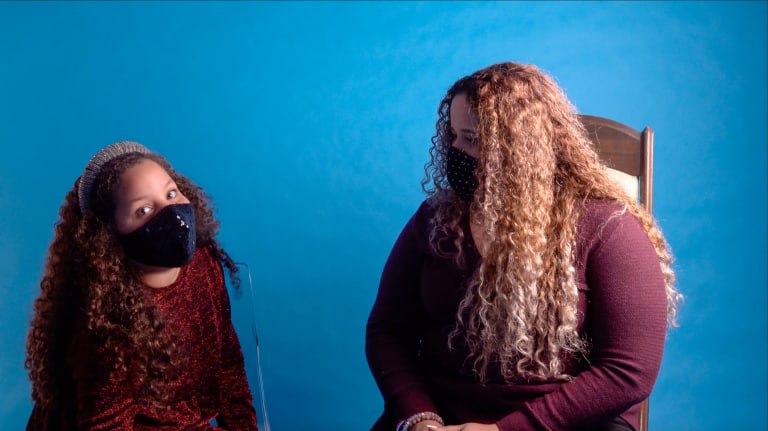 A girl with curly hair and a black mask looks at the camera as a similar-looking woman to her right looks at her. Both are seated in front of a basic blue background.
