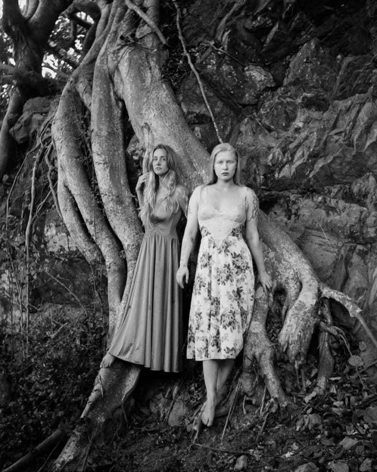 Black and white photograph of the two artists standing barefoot in front of a large tree root.