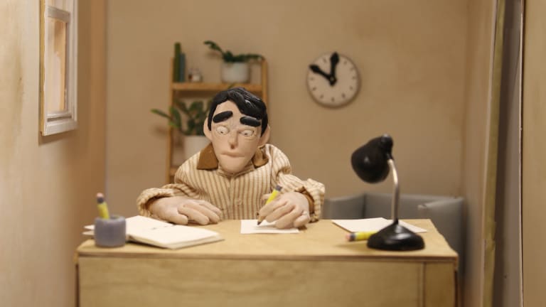 In this still image from an animation project, a puppet sits at a desk in an office.