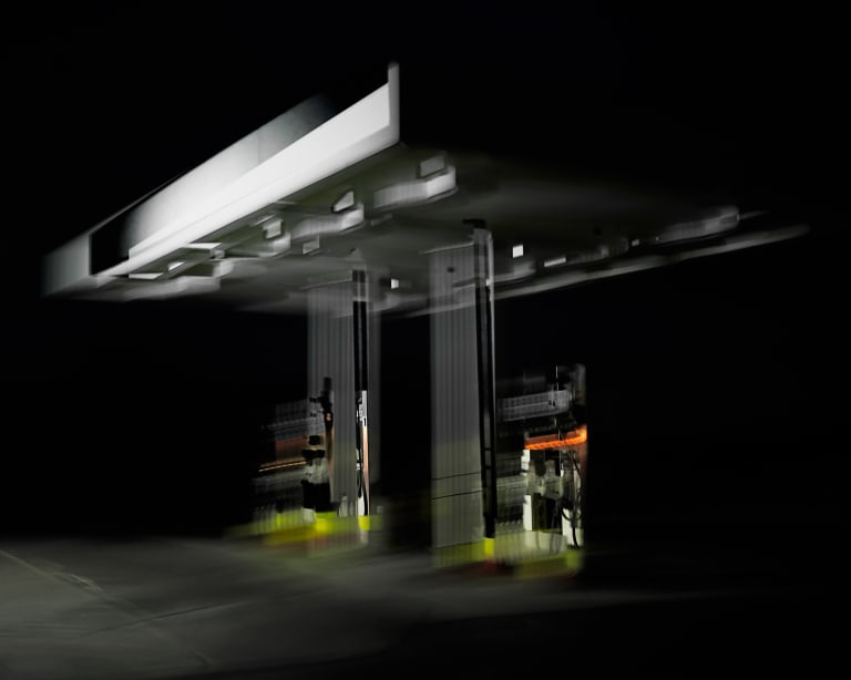 A blurry photograph of gas station pumps. The scene is lit from the left. The only color is the blurred light from the screens and the neon painted lines on the bottom lips of the concrete barriers.