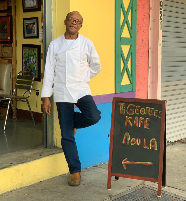 TiGeorges LaGuerre stands in front of a yellow, blue, purple, green wall outside his shop. A blackboard sign next that reads "TIGeorges Kafe" directs people inside