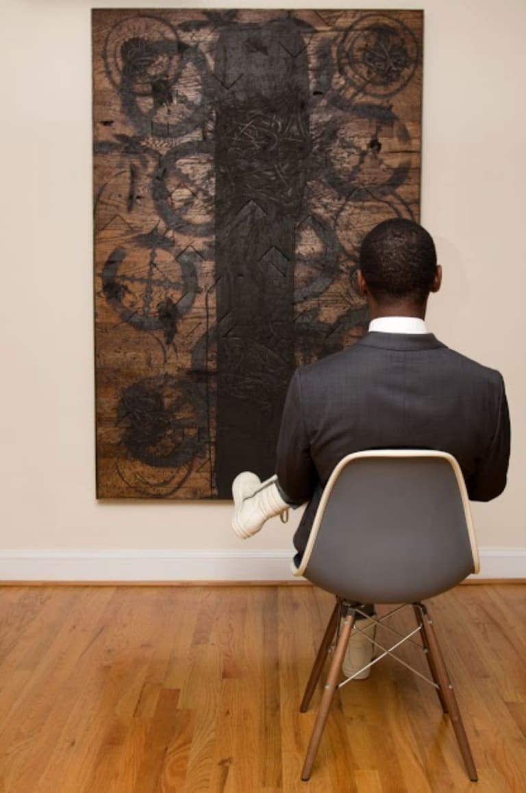 Photograph of man sitting in a chair looking at artwork, which is made of burnt wood with a large vertical line in the center and circle marks surrounding it.