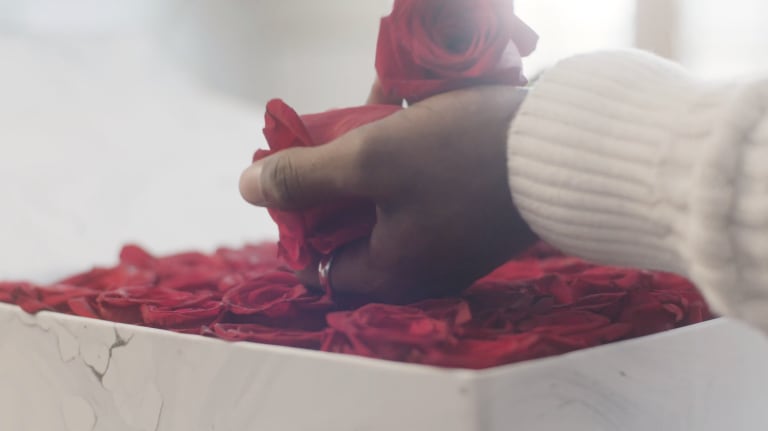 A black woman's hand holds two red roses over a container with additional roses.