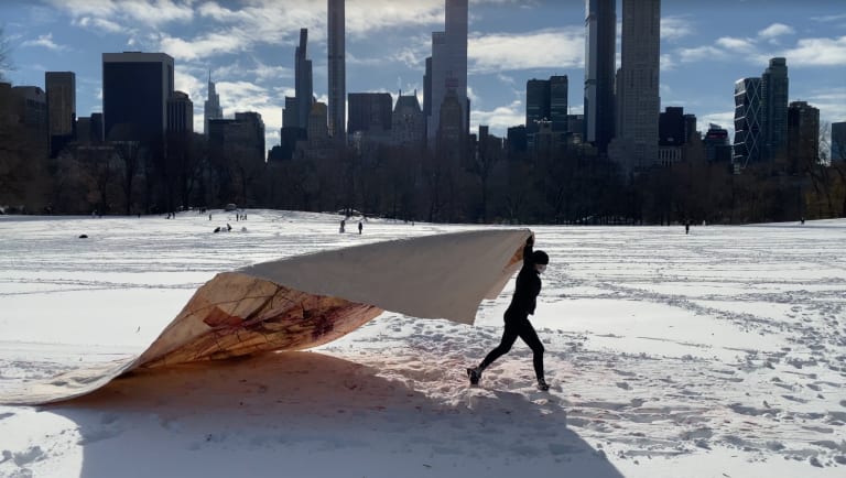 A photograph of a person dragging a large tarp behind them in Central Park on a winter's day; snow is on the ground.