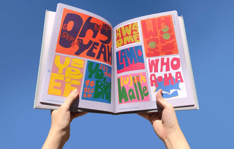 A photograph of two hands holding open a book featuring colorful, text-based artwork.