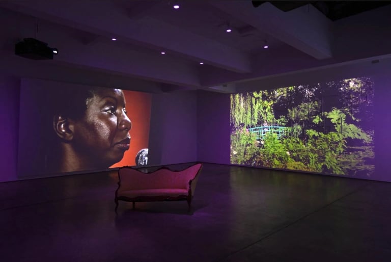 An gallery view of a multimedia installation, a black woman on the screen on the left, a green garden on the right and a sofa in the middle.