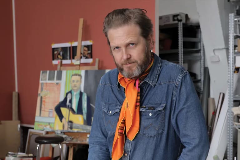 Image of Ragnar Kjartansson in his studio with a figure painting of a person playing guitar in the background