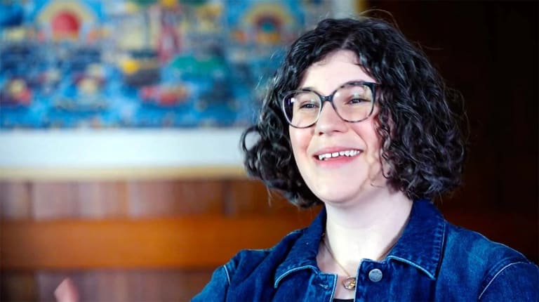 A video still of a person wearing a blue jean jacket with black curly hair and glasses who is smiling at someone offscreen.