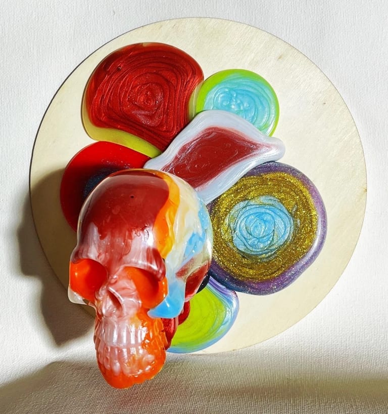 Sculpture of human skull in different colors, attached to round wooden panel with colored orbs.