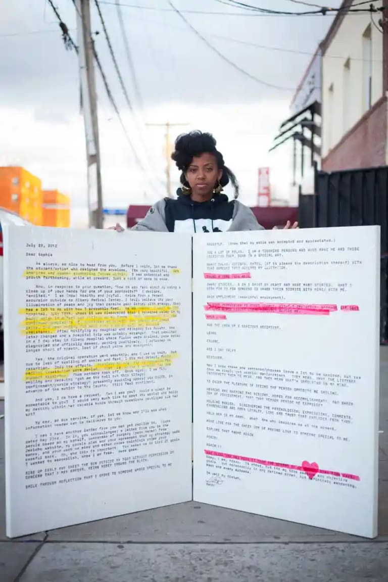 Image of artist and activist Sophia Dawson standing behind a hinged display of two drastically engarged, four-foot-tall letters wtih selected lines highlighted in yellow and pink