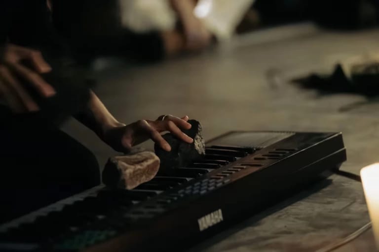 Photographic image of a hand on a keyboard. The keyboard has two rocks on it that the hand is pressing into the keys