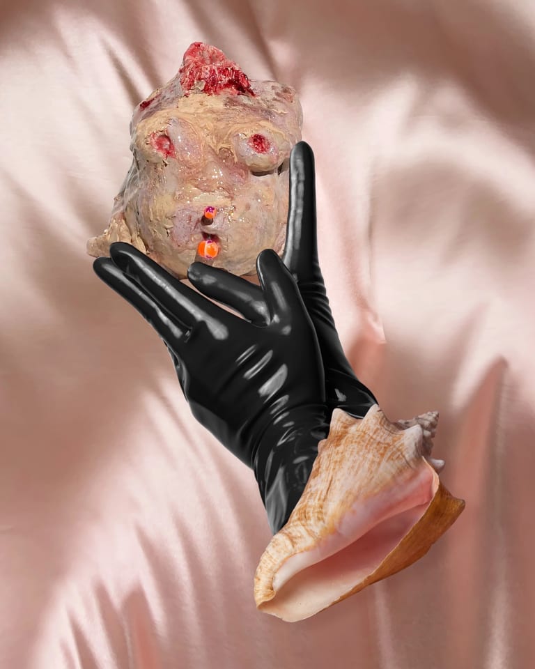 Photograph of two hands in black latex gloves coming out a shell and holding an organic amorphous shape with skin tones.
