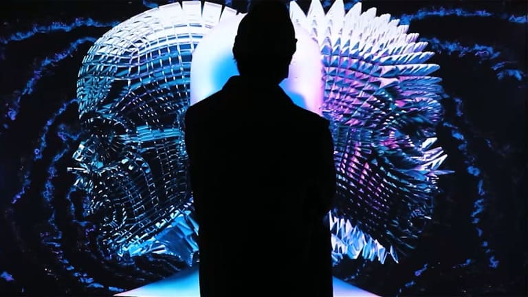 A silhouette of a person standing in front of a video projection of two abstract heads. 