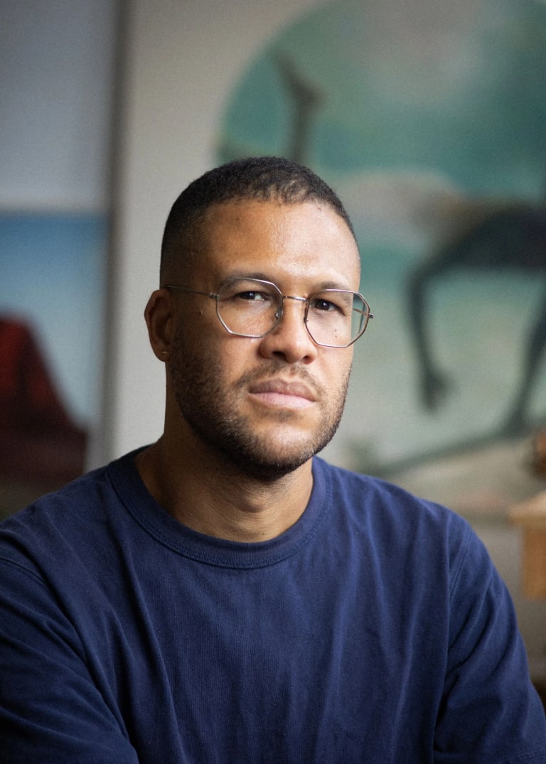 A photograph of Simon Benjamin. Simon is wearing a navy blue shirt with thin wired glasses. He is looking directly at the camera with a relaxed expression. Some of his artworks are in blurred in the background.