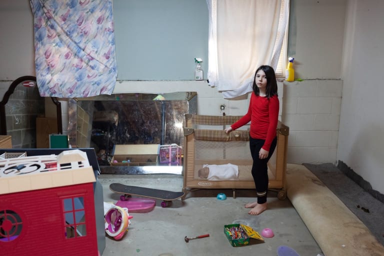 Color photograph of a girl standing next to a crib with a doll in it in a room with a dirty carpet rolled up to reveal the concrete floor, sheets over the windows and toys and mirrors scattered around the space.
