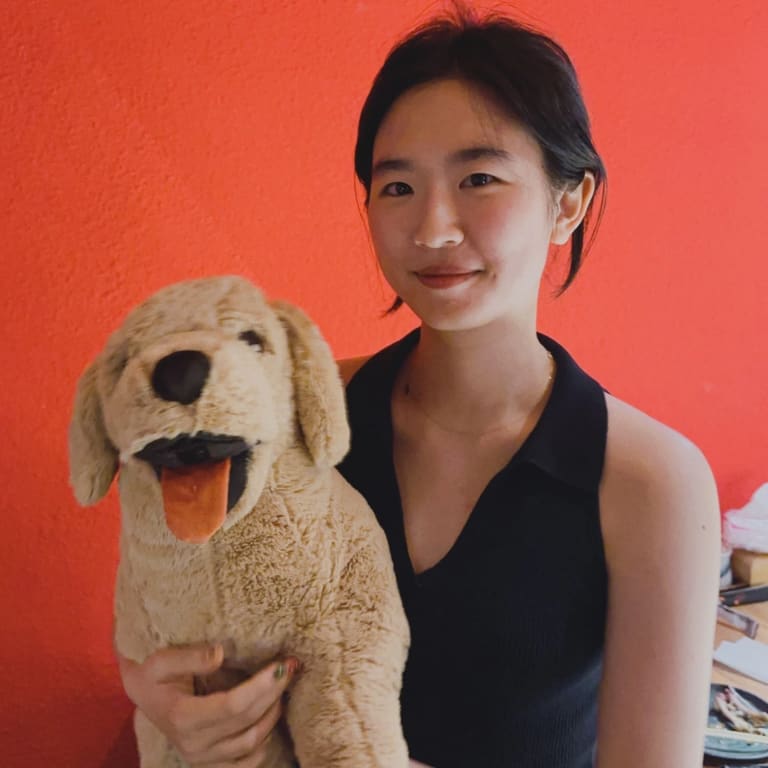 Portrait of a young woman smiling in front of an orange wall, holding a large plush doll of a dog. 