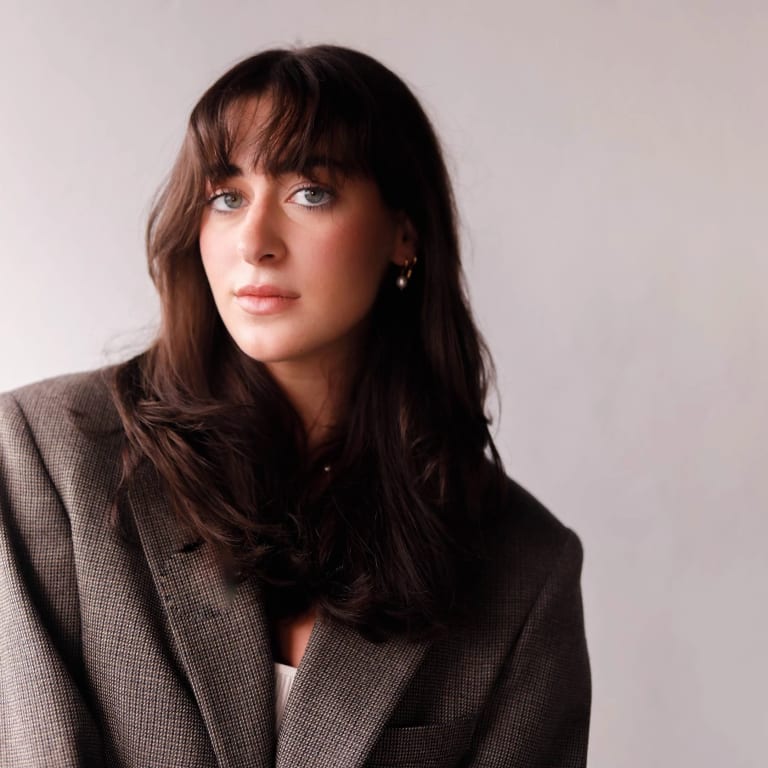 A student with brown hair and brown eyes wearing a grey suit jacket looking at the camera with a grey background.