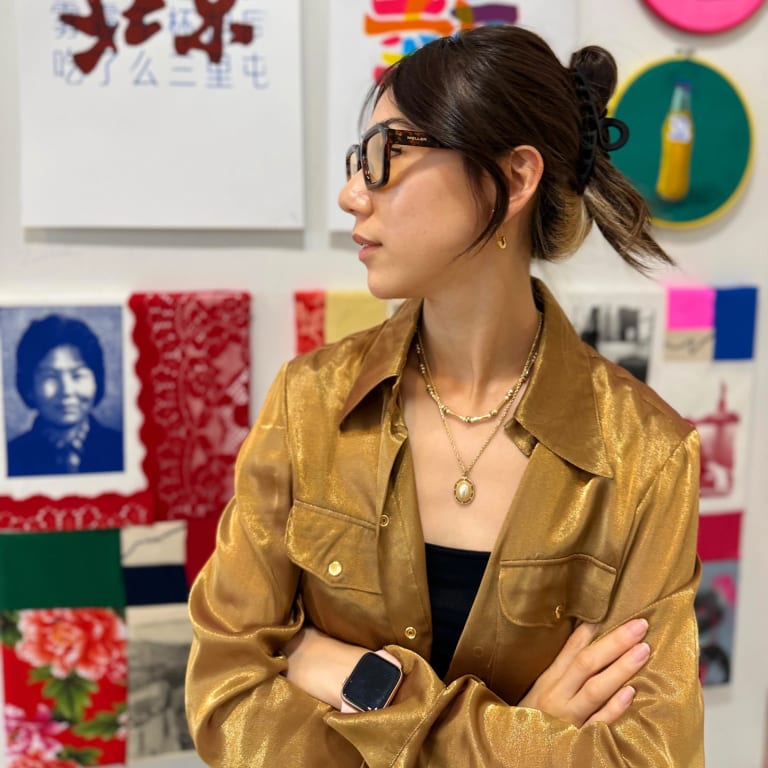 A woman in black glasses in a gold jacket standing in front of a wall with colorful art. 