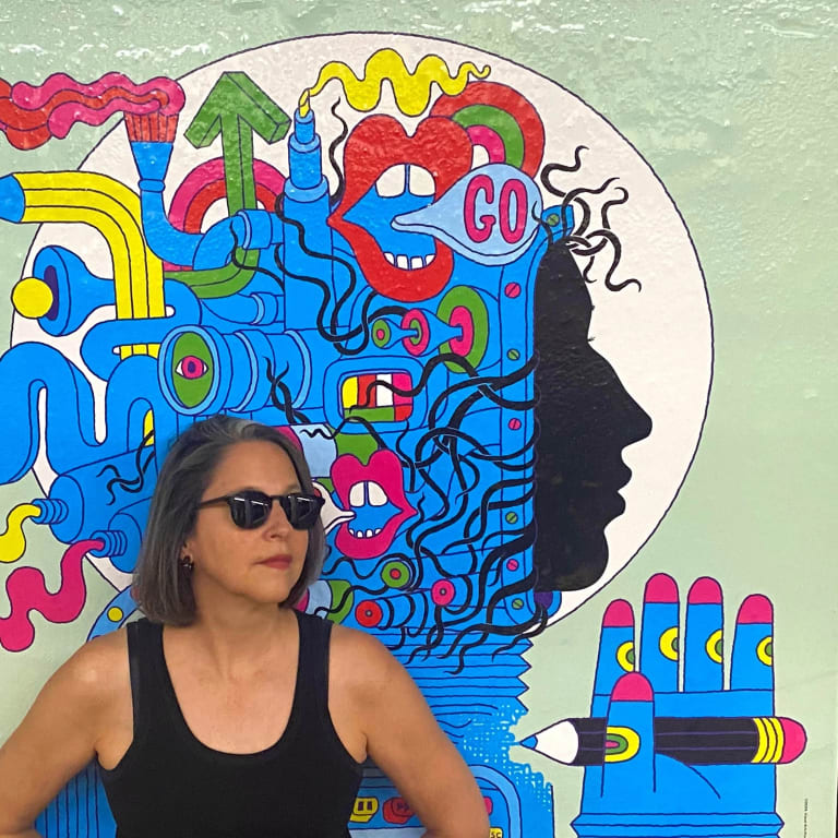 A woman in a black tank top wearing dark black sunglasses stands in front of a vibrant and colorful poster for a female astronaut in a blue and graffiti-styled space suit.