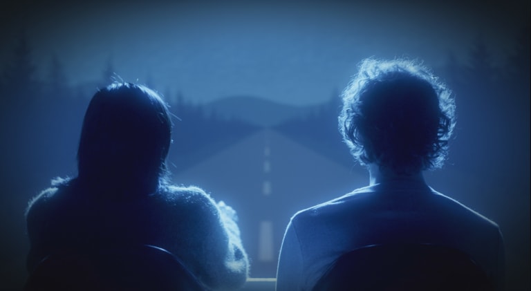 Two people sit facing away from the viewer, looking at a projected image of a long road lined with trees and a hill in the distance. A blue hue covers the image