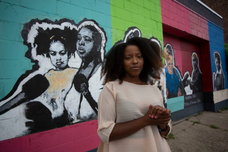 A photo of artist Sophia Dawson standing in front of a wall mural showing a black-and-white image of a Black mother and child against an aqua-colored background 