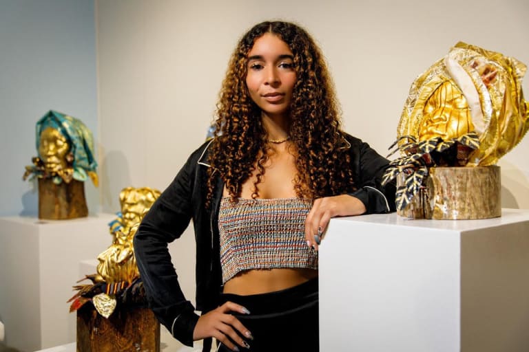 A young Black woman with long curly hair dressed in a gray tube top and a black jacket with white piping. She leans on a pedestal with a gold sculpture of a face in a hood by her left elbow, and two other pedestals with similar sculptures are behind her to her right.