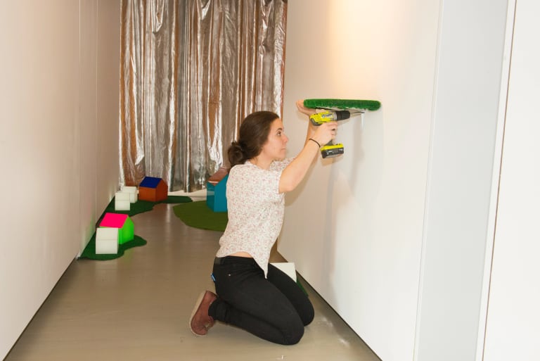 A student kneels in front of a wall in a narrow space installing work. She is holding a drill and a green object against the wall. She is in a white shirt and black jeans.