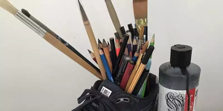 A bottle of black painting standing next to a shoe filled with pencils, pens, and paintbrushes