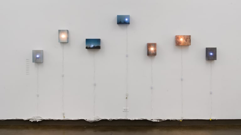 Seven small lightboxes displaying landscapes hung in a staggered row, ranging in color from deep blues to vibrant oranges.