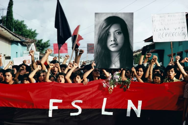 A color photograph from Nicaragua in the 1970s of protesters carrying a black and white portrait of a young woman who had been murdered and a red and black banner with the letters “F S L N” on it. 